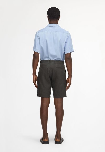 Mann von hinten, trägt ein hellblaues kurzärmeliges Hemd, dunkle Shorts und schwarze Slip-On-Sandalen vor einfarbigem weißem Hintergrund.