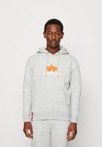 Felpa grigia con cappuccio in misto cotone, caratterizzata da un logo arancione a forma di stella sul petto, cappuccio con cordino e tasca a marsupio frontale.