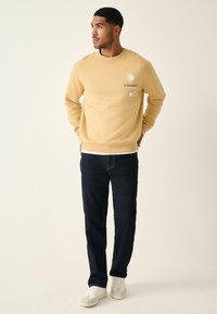 Gul sweatshirt med solgrafik på brystet, parret med mørke jeans og hvide sneakers. Enkel, afslappet design med ribbede mansjettdetaljer.