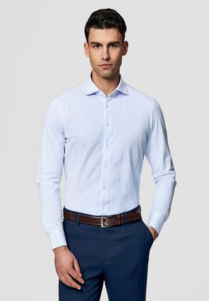 Mężczyzna w jasnoniebieskiej koszuli w paski, ciemnoniebieskich spodniach i brązowym pasku, stojący z jedną ręką w kieszeni na szarym tle.