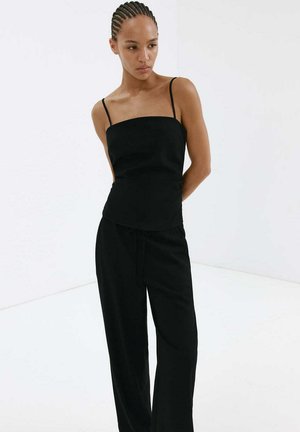 Femme aux cheveux tressés portant un haut noir sans manches avec fines bretelles et un pantalon noir ample, debout devant un fond blanc.