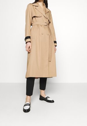 Beiger zweireihiger Trenchcoat mit Gürtel, getragen über schwarzen, verkürzten Hosen und schwarz-weißen Mokassins vor weißem Hintergrund.