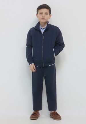 Giacca navy con zip, tasche laterali, dettagli bianchi, abbinata a pantaloni navy e scarpe marroni. Texture liscia, design casual.