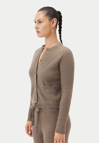 Brun stickad kofta med diamantstruktur, med rund halsringning, tre knappar och två framfickor. Stylad med en matchande midjeband.