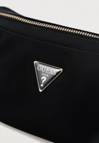 Μαύρη υφασμάτινη θήκη με υφασμάτινη υφή, που διαθέτει τριγωνικό μεταλλικό έμβλημα με την ένδειξη "GUESS" και "U.S.A." σε λευκό χρώμα και χρυσό φερμουάρ.