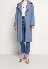 Cappotto di lana azzurro chiaro con ampio risvolto, chiusura con un bottone e polsini arrotolati, indossato sopra una maglietta bianca e abbinato a jeans aderenti e sandali con tacco.