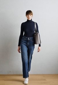 Schwarzes Rollkragenoberteil, hochgeschnittene blaue Jeans, weiße Turnschuhe und eine graue Umhängetasche. Modell läuft auf einem Holzboden vor einem hellen Hintergrund.