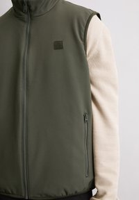 Zielona bezrękawnikowa kamizelka wykonana z gładkiego, elastycznego materiału; wyposażona w boczną kieszeń na suwak i logo na plakietce; nałożona na teksturowany kremowy top.