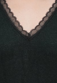 Nahaufnahme eines schwarzen Strickgewands mit einem V-Ausschnitt, der mit zarter schwarzer Spitze verziert ist, vor heller Haut.