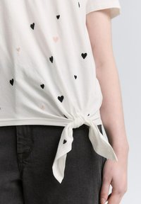 Biała koszulka z czarnymi i różowymi wzorami serc, związana z boku. Wykonana z miękkiej bawełny, o luźnym kroju. Połączona z ciemnymi dżinsami.