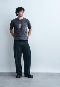 Νεαρός με σγουρά μαλλιά που φοράει ξεθωριασμένο μαύρο μπλουζάκι με σχέδιο, άνετο σκούρο τζιν και μαύρα χοντρά παπούτσια, που στέκεται μπροστά σε έναν απλό τοίχο.