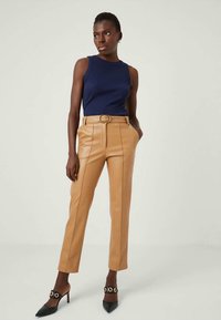 Kvinna står med händerna i fickorna, iklädd en marinblå ärmlös topp, beige bältade läderbyxor och svarta spetsiga klackskor med guldaccenter.