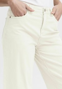Light beige trousers made of cotton, featuring a straight-leg design, front pockets, and stitched seams. Close-up view of the waistband area.