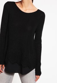 Svart stickad tröja med långa ärmar, med en scalloped spetskant. Tyget är texturerat; passformen är avslappnad och något oversized.