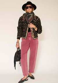 Veste noire à motif floral avec bordure dorée, col roulé noir, jean rose taille haute, sac à main noir et chapeau noir. Lunettes de soleil noires.