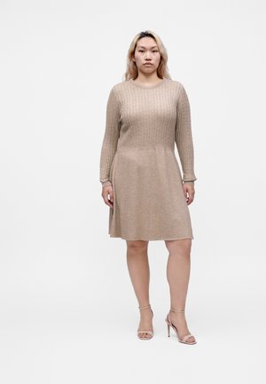 Femme debout face à l’avant, portant une robe en maille beige à manches longues et des sandales à talons hauts couleur nude sur un fond blanc uni.