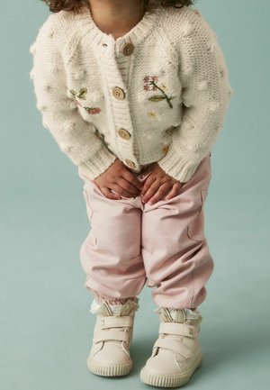 Maluch ubrany w kremowy sweter na drutach z kwiatową haftem, różowe spodnie i kremowe buty z uszami królika, stoi z rękami na kolanach.