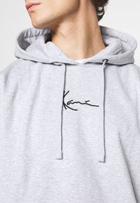 Jeune personne portant un sweat à capuche gris clair avec un logo noir de style manuscrit sur la poitrine, tête tournée vers la gauche.