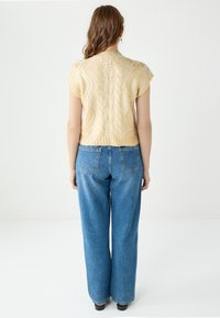 Gehaakte gele trui met een kabelpatroon en korte mouwen, gecombineerd met hoge blauwe wijde jeans. Witte achtergrond.