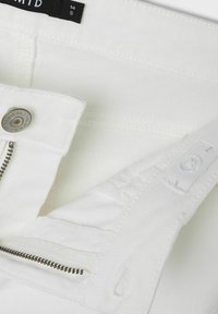 Közelkép fehér farmernadrág cipzárjáról, "Denim Brand Original" felirattal ellátott fémbotról, és az állítható derék gomblyuk részletéről.