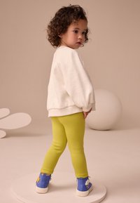 Weiß gefleckter Pullover, gelbe Leggings, blaue High-Top-Sneaker mit Cartoon-Akzenten, Kind schaut nach hinten, hellbeiger Hintergrund.