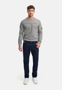Homme portant un sweat-shirt gris avec le texte « INDICODE JEANS » et un jean bleu foncé, debout devant un fond blanc uni.