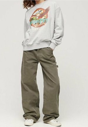 Persoon draagt een grijze sweatshirt met arendgrafiek en groene wijde broekspijpen, handen in de zakken, staand met witte sneakers.