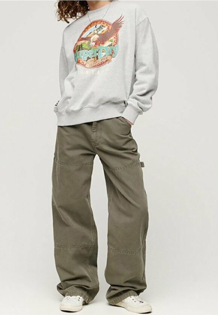 Personne portant un sweat-shirt gris avec un motif d’aigle et un pantalon vert à larges jambes, mains dans les poches, debout avec des baskets blanches.