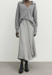 Personne portant un pull en V gris, une jupe drapée gris clair avec une ceinture, des chaussures noires à talons pointus, se tenant devant un mur blanc uni.