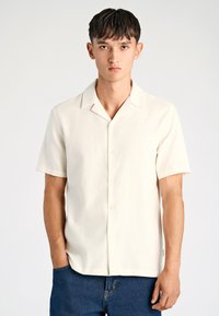 Camisa de manga corta en crema claro, con una textura suave, cuello abierto y botones en la parte delantera. Combinada con jeans de mezclilla oscuros.