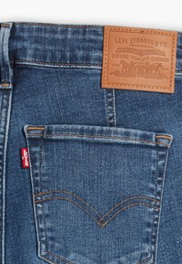 Poche arrière d'un jean Levi's en denim bleu avec des coutures orange, une étiquette Levi's rouge et un patch en cuir brun de la marque au-dessus de la poche.