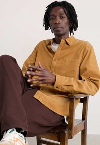 Bruine corduroy shirt met knopenlijst, gecombineerd met donkerbruine broek. Model zit op een houten stoel met de handen gevouwen.