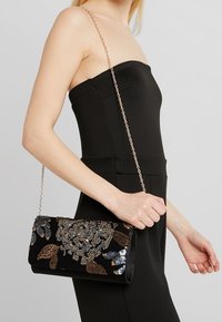Svart clutch med dekorativa paljetter och broderimönster i guld, silver och brunt, med en avtagbar kedjeskarp.