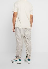 Person, der ein beiges Kurzarmhemd, eine hellbeige Jogginghose und weiße FILA-Sneaker trägt, steht vor einem einfarbigen weißen Hintergrund.
