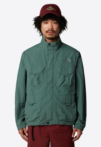 Grüne, leichte Jacke mit hohem Kragen, mehreren Fronttaschen und elastischen Bündchen. Mit Logo-Stickerei auf der linken Brust.