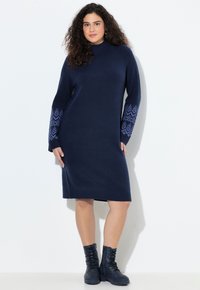 Abito a maglia a maniche lunghe di colore blu navy con colletto alto e polsini decorati; abbinato a stivali alla caviglia neri con lacci. Texture liscia e vestibilità comoda.