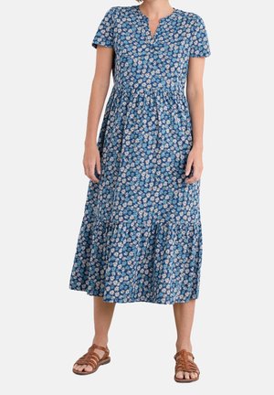 Woman wearing a blue midi dress with white daisy patterns, short sleeves, v-neckline, and brown flat sandals, standing on a plain background.