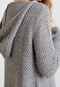 Grå stickad cardigan med ett strukturerat mönster, huva och detaljer med ribbade muddar. Materialet verkar mjukt och lätt.