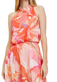 Kvinna i ärmlös klänning med hög hals och abstrakt blommönster i nyanser av orange, rosa och vitt.