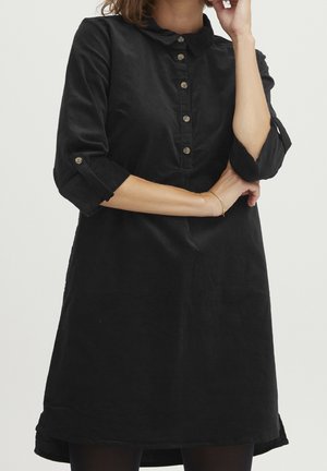 Femme portant une robe noire à manches longues avec des boutons sur la poitrine et des manches retroussées jusqu'aux coudes, debout les bras croisés.