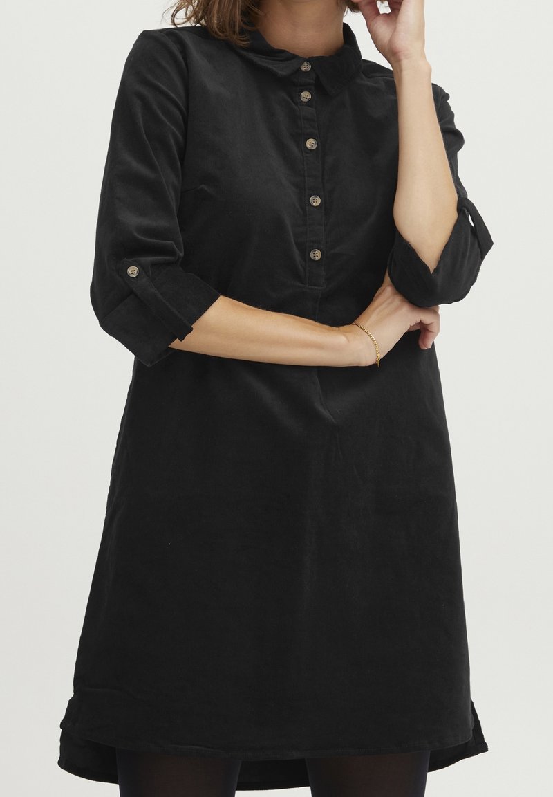 Femme portant une robe noire à manches longues avec des boutons sur la poitrine et des manches retroussées jusqu'aux coudes, debout les bras croisés.