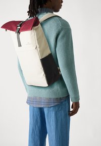Rucsac cu un capac de culoare bordo, corp crem și fund negru. Prezintă bretele reglabile și un design elegant, minimalist.