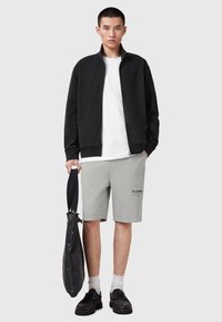 Zwarte ritsjack, witte T-shirt, grijze shorts met logo, zwarte leren tas, zwarte schoenen en witte sokken. Casual sportieve look.