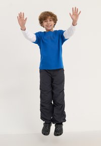 Blue short-sleeve shirt over a white long-sleeve shirt, black loose pants, and black clogs, with arms raised in a jumping pose.