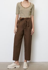 Pruhovaná olivově zelená a krémová blůza s kulatým výstřihem, spárovaná s hnědými kalhotami s vysokým pasem a uvázaným páskem. Béžové dřeváky s tlustými podrážkami.