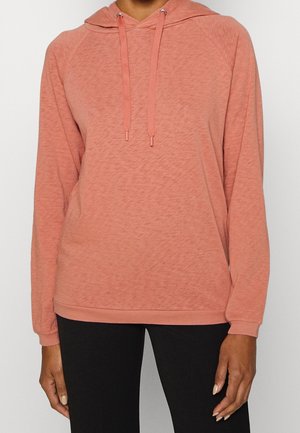 Vrouw draagt een koraalroze hoodie met lange mouwen en trekkoorden en zwarte broek, staand tegen een effen witte achtergrond.