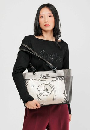 Silberne und schwarze Handtasche mit grafischem Gesichtdesign, aus Leder, mit doppeltem Tragegurt und dem eingravierten Markennamen "Anekke" auf der Vorderseite.