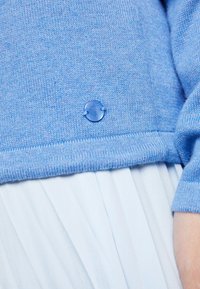 Ljust blått stickat tröja med en rund blå knapp i nederkan. Mjuk textur med ribbade ärmavslut och en något avslappnad passform.