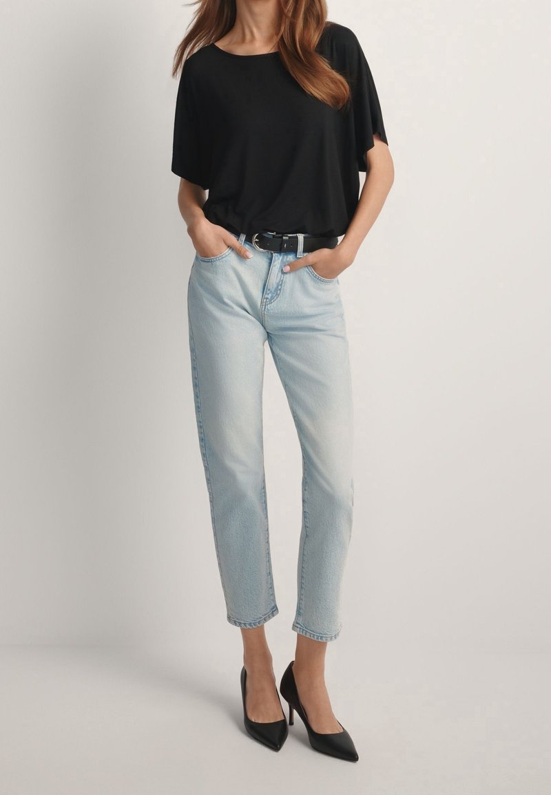 Femme portant un haut noir à manches courtes, un jean bleu clair taille haute avec une ceinture noire, et des escarpins noirs pointus, debout les mains dans les poches.