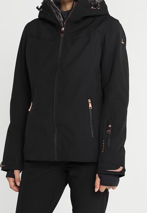 Zwarte winterjas met capuchon afgezet met bont, meerdere ritszakken en verlengde polsboorden, gedragen door een persoon die zwarte broek draagt.